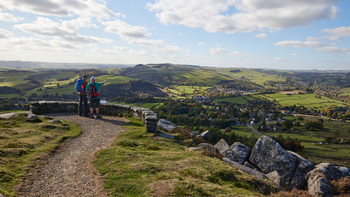 Peak district view This landscape photograph captures a panoramic afternoon view of Curbar Edge in the Peak District, Derbyshire, United Kingdom, during the autumn season. The image showcases the sweeping rural scenery of the region, with expansive green fields and gently rolling hills characteristic of nature in the Peak District. A pair of hikers with backpacks stands at a viewpoint overlooking the valley, adding a sense of scale to the vast surroundings. In the foreground, rocks and grassy paths indicate the rugged terrain typical of Curbar Edge, while in the distance, scattered villages and wooded areas highlight the rural charm of Derbyshire. The scene is well-lit by soft sunlight filtering through a partly cloudy sky, emphasizing the vibrant autumnal colors and providing clear visibility across the landscape.